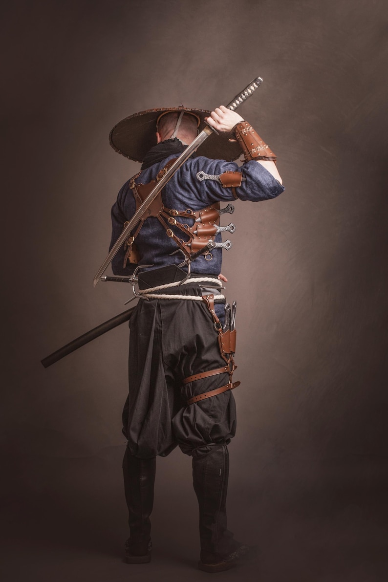 May include: A person wearing a blue shirt and black pants with a wide-brimmed hat, holding a sword over their shoulder. They are wearing a leather harness with multiple knives attached.