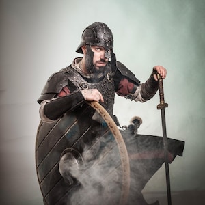 K&ouml;nnte beinhalten: Eine Person in voller R&uuml;stung, einschlie&szlig;lich Helm, Schulterpolstern und Brustplatte, h&auml;lt einen Schild und ein Schwert. Der Schild ist rund und aus Holz, und das Schwert ist lang und gerade. Die R&uuml;stung ist schwarz und detailliert, mit roten Akzenten. Das Bild hat einen rauchigen Effekt.