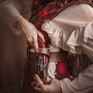 Puede incluir: Una mujer con un traje rojo y blanco con un cintur&oacute;n y una funda de cuero marr&oacute;n. Una pistola negra y plateada est&aacute; en la funda. La mujer est&aacute; sujetando la funda con su mano derecha.