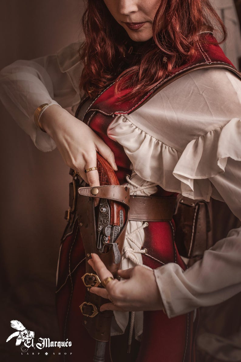 Puede incluir: Una mujer con un traje rojo y blanco con un cintur&oacute;n y una funda de cuero marr&oacute;n. Una pistola negra y plateada est&aacute; en la funda. La mujer est&aacute; sujetando la funda con su mano derecha.
