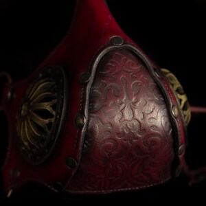 May include: A red leather mask with intricate gold metal accents and a floral design. The mask has two straps that attach to the back of the head.