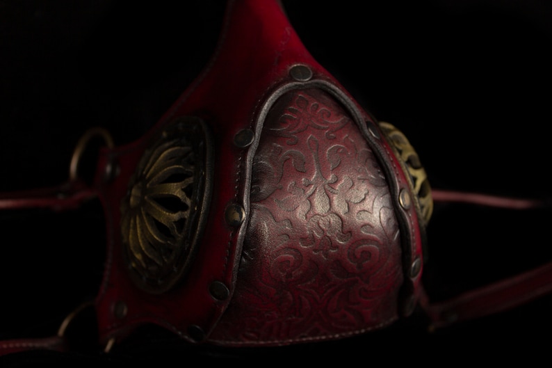May include: A red leather mask with intricate gold metal accents and a floral design. The mask has two straps that attach to the back of the head.