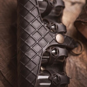 Peut inclure: Brassard en cuir noir avec un motif en losanges et des lani&egrave;res d&eacute;coratives. Le brassard a une fermeture &agrave; bouton en laiton et est con&ccedil;u pour la protection. C'est une pi&egrave;ce d'armure.