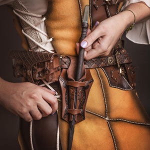 Peut inclure: Une ceinture en cuir marron avec un &eacute;tui d&eacute;coratif contenant une baguette. La ceinture est port&eacute;e sur une tunique en cuir jaune et marron. La baguette a un manche fonc&eacute; et une pointe en cristal transparent. La tenue est d&eacute;taill&eacute;e avec des coutures et des boucles.
