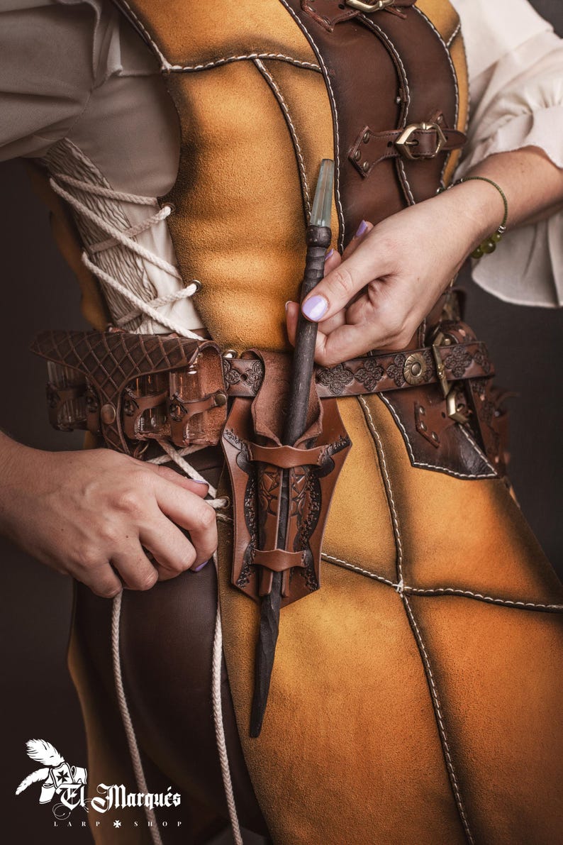 Peut inclure: Une ceinture en cuir marron avec un &eacute;tui d&eacute;coratif contenant une baguette. La ceinture est port&eacute;e sur une tunique en cuir jaune et marron. La baguette a un manche fonc&eacute; et une pointe en cristal transparent. La tenue est d&eacute;taill&eacute;e avec des coutures et des boucles.