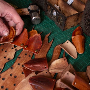 May include: A close-up of a wooden board with holes and leather pieces being placed on top. The leather pieces are various shades of brown and are cut into different shapes. The board is on a green cutting mat.