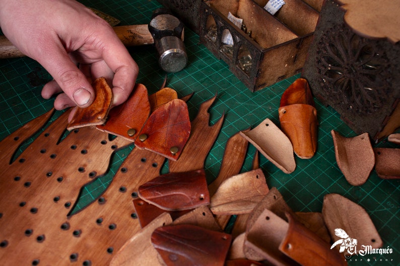 May include: A close-up of a wooden board with holes and leather pieces being placed on top. The leather pieces are various shades of brown and are cut into different shapes. The board is on a green cutting mat.