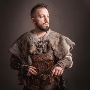 May include: A person wearing a brown leather armor with intricate carvings, a fur-lined cloak, and a sword. The armor is decorated with a belt and buckles. The person is standing against a gray background.