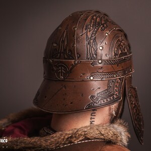 K&ouml;nnte beinhalten: Brauner Lederhelm mit aufwendigen gepr&auml;gten Designs, Nieten und einem Visier. Der Helm weist dekorative Muster auf und ist Teil eines Kost&uuml;ms oder einer historischen Nachstellung. Das Bild zeigt die R&uuml;ckseite des Helms.