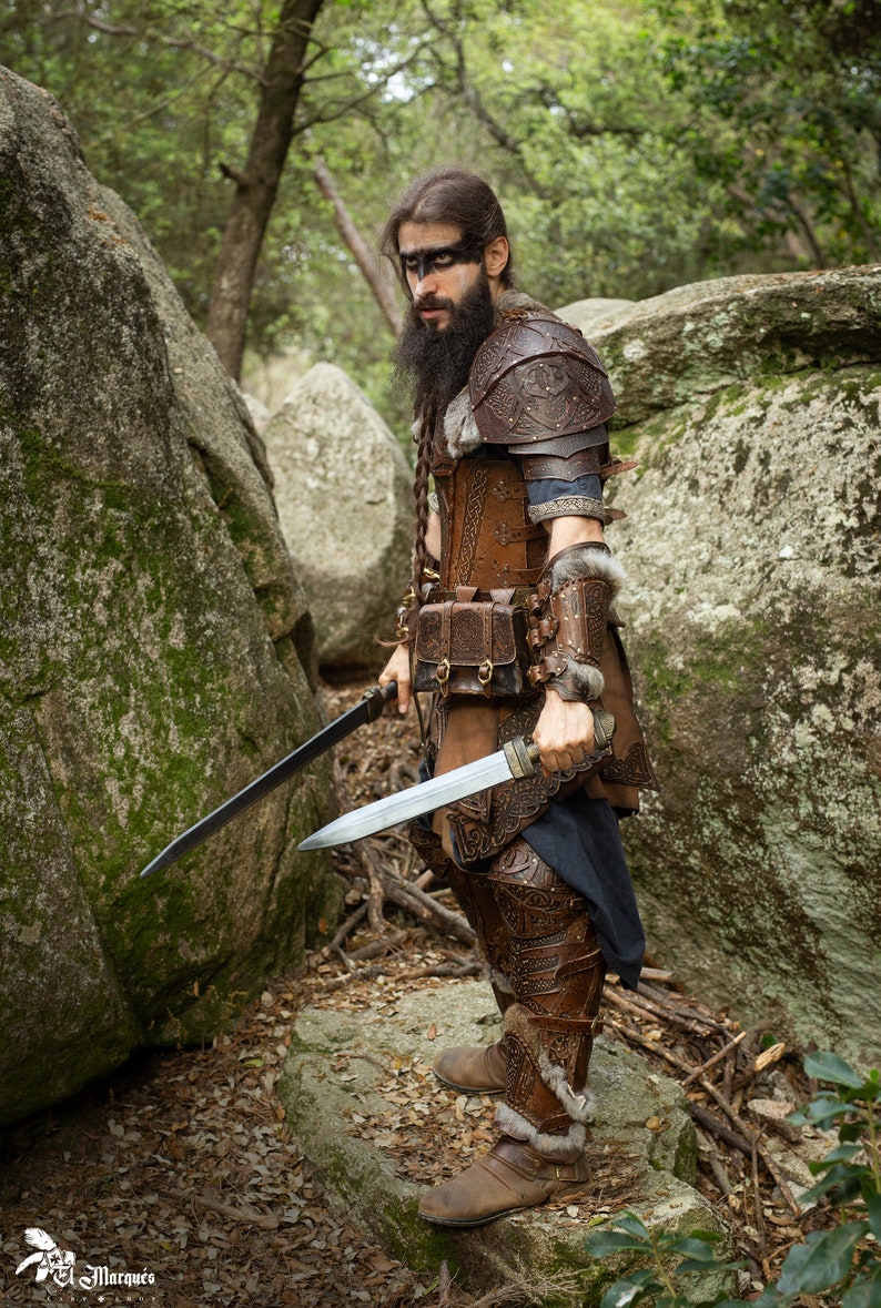 May include: A man dressed in leather armor and fur holds two swords in a forest setting. He has a beard and dark paint on his face.