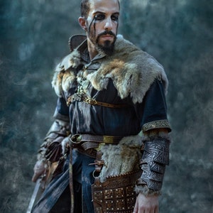 May include: A man dressed in a dark blue tunic with fur trim, leather armor, and a sword. He has dark hair and a beard, and his face is painted with black markings.