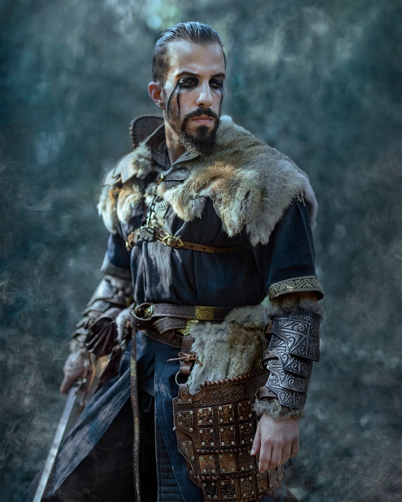 May include: A man dressed in a dark blue tunic with fur trim, leather armor, and a sword. He has dark hair and a beard, and his face is painted with black markings.