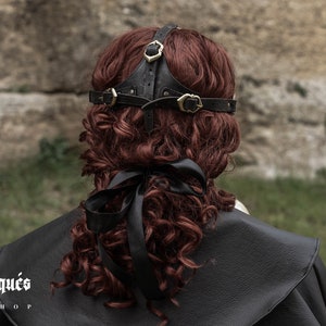 May include: Black leather headband with gold metal accents. The headband is worn on a person with long, curly red hair.