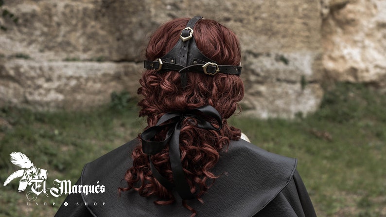 May include: Black leather headband with gold metal accents. The headband is worn on a person with long, curly red hair.
