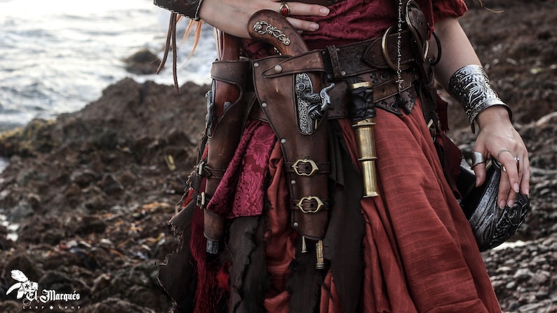 Puede incluir: Una mujer con un vestido rojo con un cintur&oacute;n de cuero marr&oacute;n y dos pistolas en fundas en sus caderas. Las pistolas est&aacute;n en fundas de cuero marr&oacute;n con detalles met&aacute;licos. La mujer tambi&eacute;n lleva una pulsera de plata en su mu&ntilde;eca derecha.