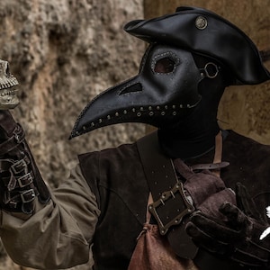May include: A person wearing a black plague doctor mask and hat holds a white skull in their gloved hand. The person is wearing a brown leather jacket and a black shirt. The background is a stone wall.