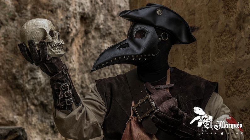 May include: A person wearing a black plague doctor mask and hat holds a white skull in their gloved hand. The person is wearing a brown leather jacket and a black shirt. The background is a stone wall.