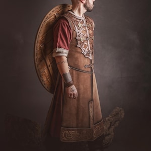 Peut inclure: Une personne portant une tunique en cuir marron avec des coutures d&eacute;coratives et un sous-v&ecirc;tement rouge, portant un bouclier en bois. La tenue rappelle les v&ecirc;tements historiques ou fantastiques. L'image comprend le logo de "El Marqu&eacute;s LARP Shop".