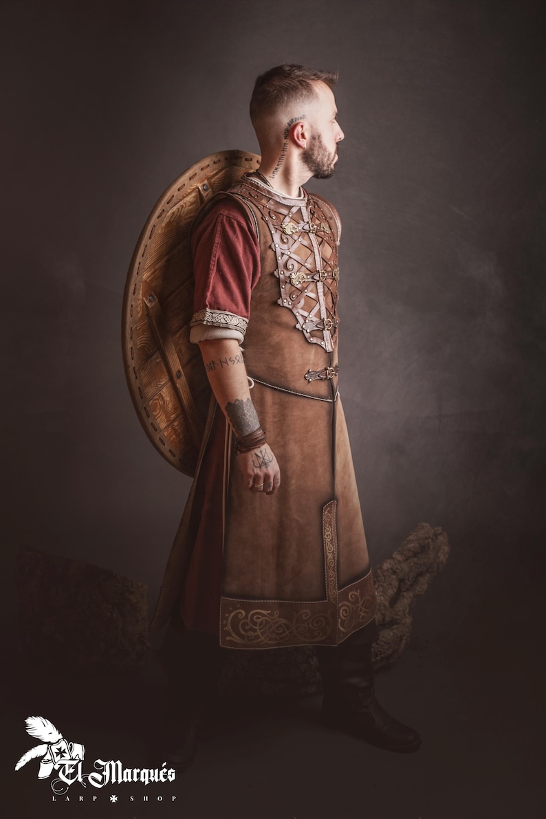 Peut inclure: Une personne portant une tunique en cuir marron avec des coutures d&eacute;coratives et un sous-v&ecirc;tement rouge, portant un bouclier en bois. La tenue rappelle les v&ecirc;tements historiques ou fantastiques. L'image comprend le logo de "El Marqu&eacute;s LARP Shop".