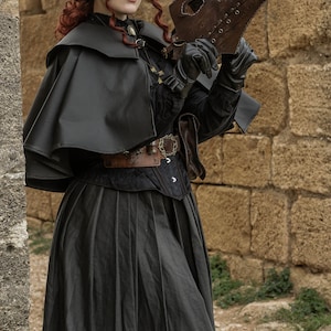 May include: A woman in a black costume with a long black skirt, a black cape, and a black hat with a feather. She is holding a brown leather plague doctor mask with a long beak. The mask has metal rivets and a leather strap. The woman is standing in front of a stone wall.