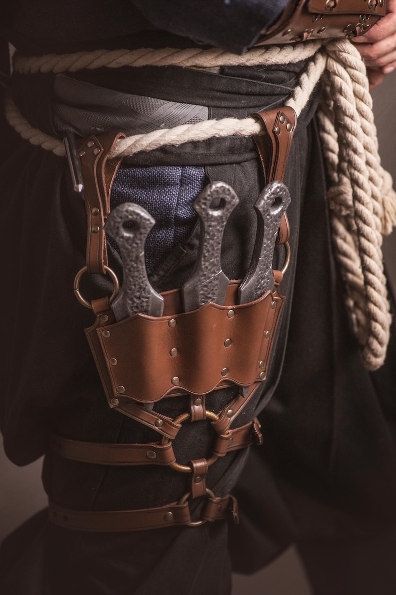 May include: A brown leather sheath with three silver throwing knives attached to a brown leather belt with metal buckles and rings.