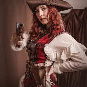 Puede incluir: Una mujer vestida con un traje de pirata rojo y marr&oacute;n sostiene una pistola negra en su mano derecha. Lleva un sombrero de cuero marr&oacute;n con un dise&ntilde;o de br&uacute;jula, una blusa blanca con volantes y un chaleco de cuero rojo con correas y hebillas de cuero marr&oacute;n. Tambi&eacute;n lleva un cintur&oacute;n de cuero marr&oacute;n con una funda para una pistola.