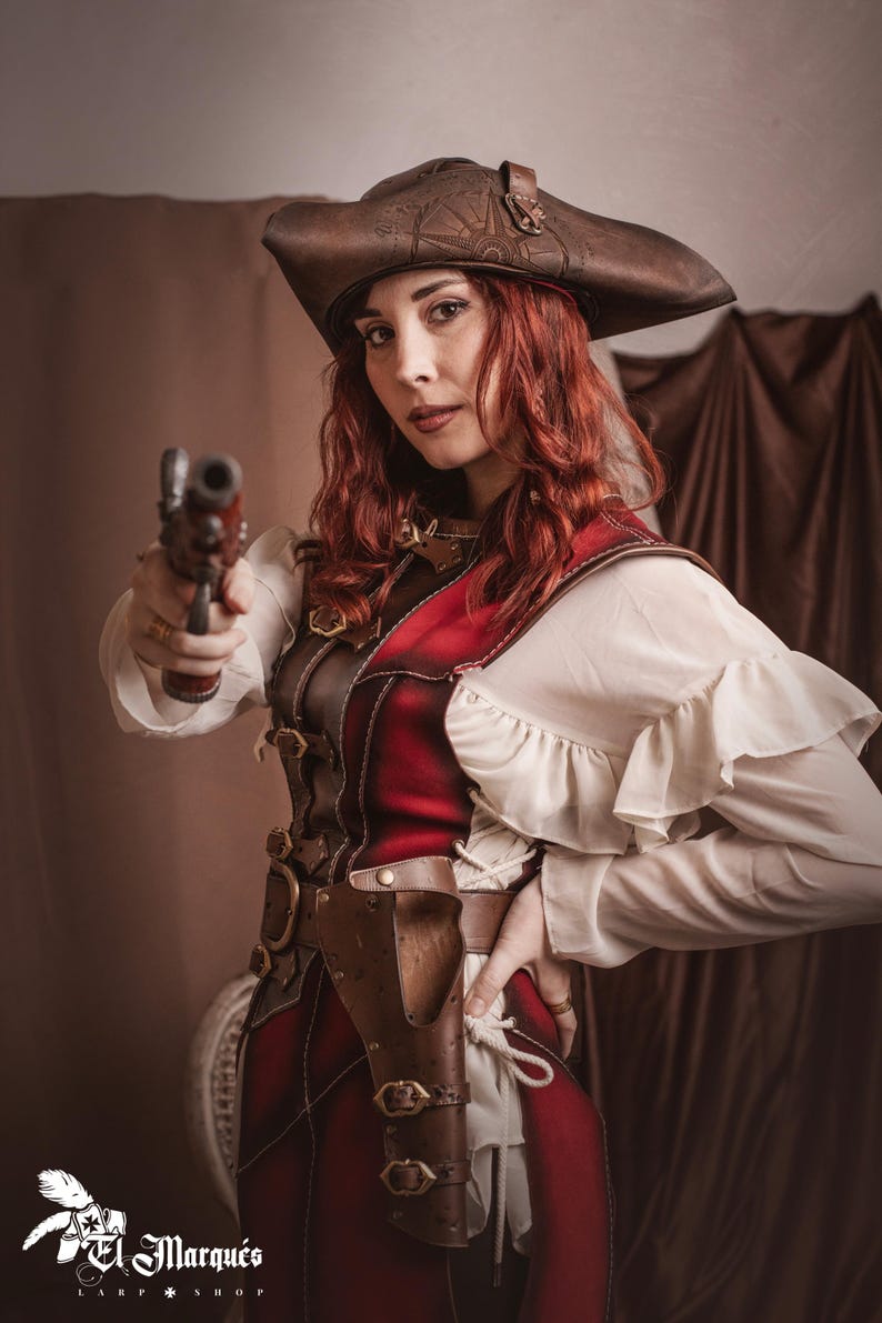 Puede incluir: Una mujer vestida con un traje de pirata rojo y marr&oacute;n sostiene una pistola negra en su mano derecha. Lleva un sombrero de cuero marr&oacute;n con un dise&ntilde;o de br&uacute;jula, una blusa blanca con volantes y un chaleco de cuero rojo con correas y hebillas de cuero marr&oacute;n. Tambi&eacute;n lleva un cintur&oacute;n de cuero marr&oacute;n con una funda para una pistola.