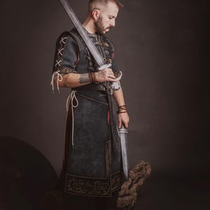 Peut inclure: Un costume m&eacute;di&eacute;val gris fonc&eacute; avec des d&eacute;tails dor&eacute;s, une longue &eacute;p&eacute;e et un poignard. Le costume pr&eacute;sente des coutures complexes et des d&eacute;tails en cuir. L'image est sur fond sombre, mettant en valeur les d&eacute;tails du costume et des armes.