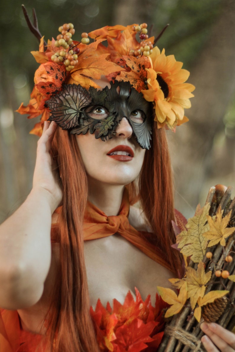 May include: A woman wearing a leafy, autumn-themed mask with sunflowers and berries. She has long red hair and is wearing a brown and orange outfit with a leafy bodice.