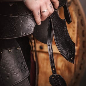 K&ouml;nnte beinhalten: Nahaufnahme einer schwarzen Lederr&uuml;stung mit Nieten und Riemen. Die R&uuml;stung wird von einer Hand mit T&auml;towierungen und einem Silberring gehalten. Im Hintergrund ist ein Holzschild zu sehen. Das Bild hat ein mittelalterliches oder Fantasy-Thema.