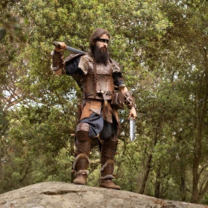 May include: A man dressed in a brown leather costume with a sword and a dagger, standing on a rock in a forest.
