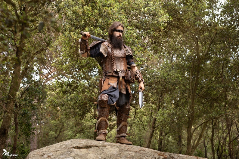 May include: A man dressed in a brown leather costume with a sword and a dagger, standing on a rock in a forest.