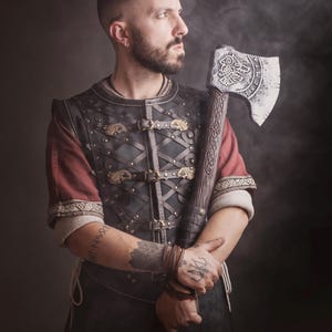 Peut inclure: Une personne v&ecirc;tue d'une tenue de style viking, tenant une grande hache orn&eacute;e. La tenue comprend un gilet en cuir avec des fermoirs dor&eacute;s et des manches rouges. La t&ecirc;te de la hache est argent&eacute;e avec des gravures complexes. Fond sombre et enfum&eacute;.