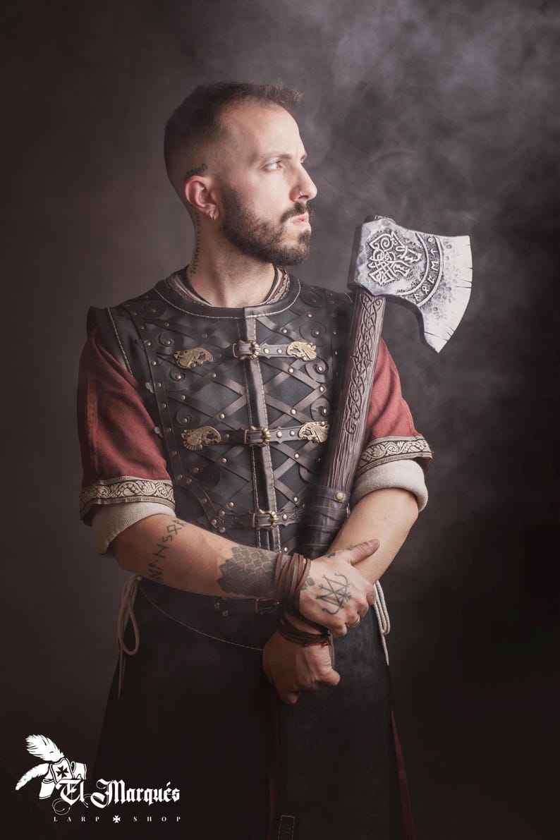 Peut inclure: Une personne v&ecirc;tue d'une tenue de style viking, tenant une grande hache orn&eacute;e. La tenue comprend un gilet en cuir avec des fermoirs dor&eacute;s et des manches rouges. La t&ecirc;te de la hache est argent&eacute;e avec des gravures complexes. Fond sombre et enfum&eacute;.