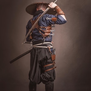 May include: A person wearing a blue shirt and black pants with a wide-brimmed hat, holding a sword over their shoulder. They are wearing a leather harness with multiple knives attached.
