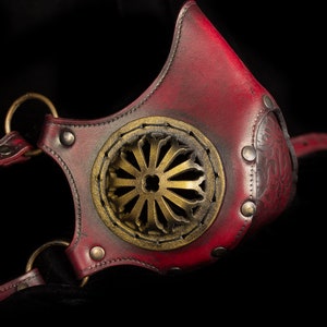 May include: A red leather mask with a gold metal design and straps. The mask has a circular opening in the front with a decorative metal design.