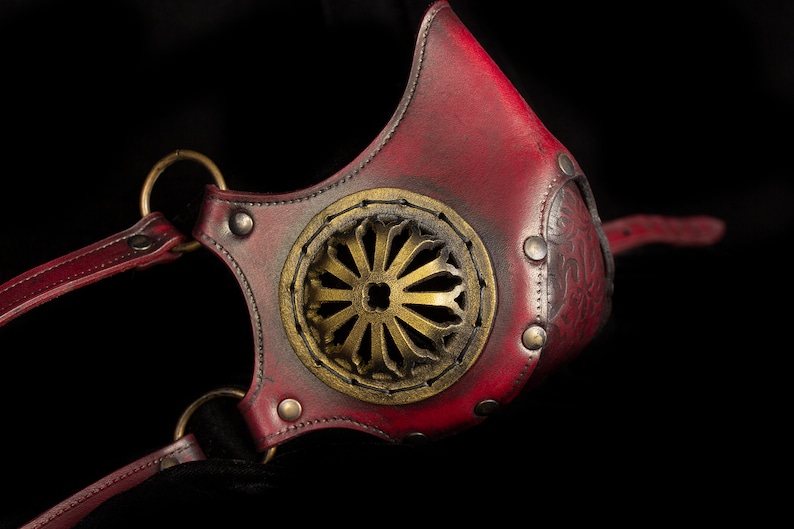 May include: A red leather mask with a gold metal design and straps. The mask has a circular opening in the front with a decorative metal design.