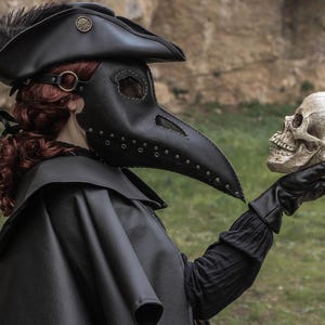 May include: A person wearing a black leather plague doctor mask with a long beak and a black hat with a feather. They are holding a white skull in their gloved hand.