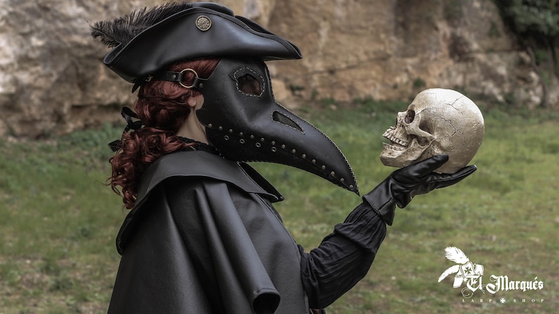 May include: A person wearing a black leather plague doctor mask with a long beak and a black hat with a feather. They are holding a white skull in their gloved hand.