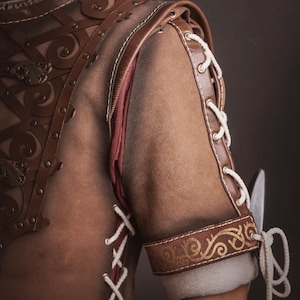 Peut inclure: Gros plan sur une armure en cuir marron avec des coutures d&eacute;coratives et des accents dor&eacute;s. L'&eacute;paule et le bras pr&eacute;sentent des motifs complexes et des d&eacute;tails de la&ccedil;age. L'armure est probablement destin&eacute;e au cosplay ou &agrave; la reconstitution historique.