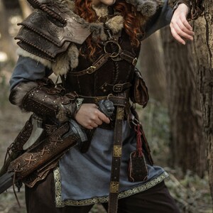 May include: A woman in a forest setting, wearing a brown leather and fur costume with a sword strapped to her side. She has red hair and blue face paint.