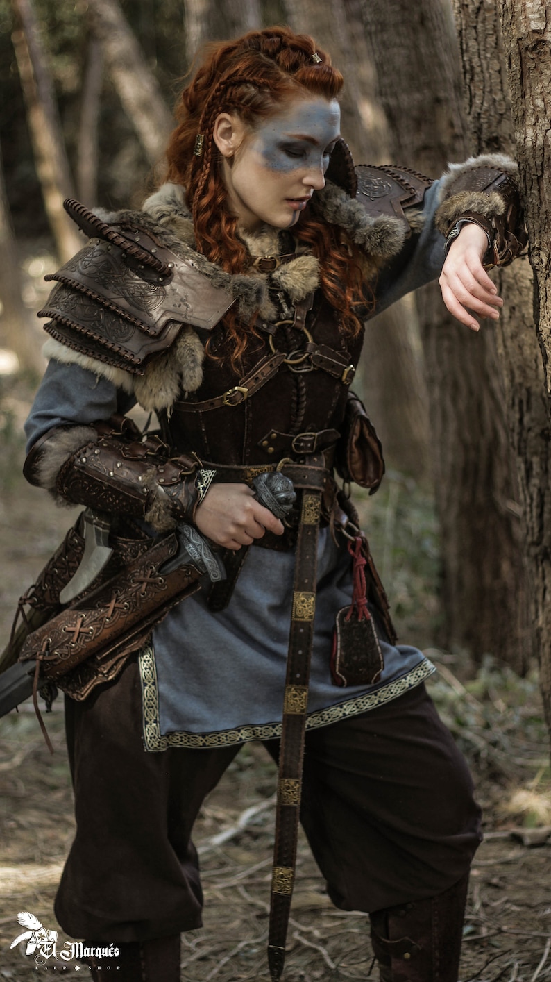 May include: A woman in a forest setting, wearing a brown leather and fur costume with a sword strapped to her side. She has red hair and blue face paint.