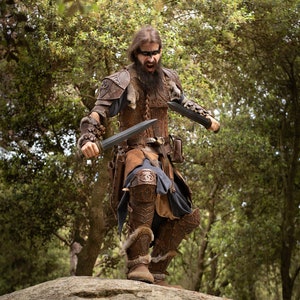 May include: A man wearing a leather costume with fur accents and holding two swords. He is standing on a rock in a forest setting.