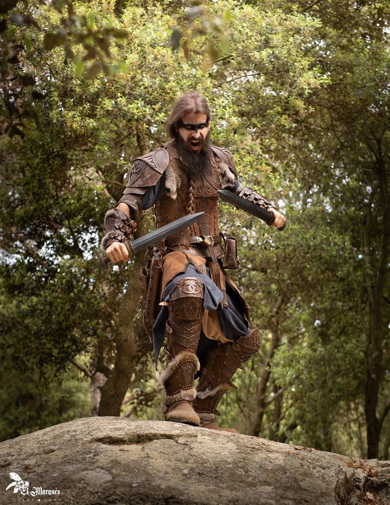 May include: A man wearing a leather costume with fur accents and holding two swords. He is standing on a rock in a forest setting.