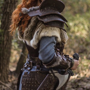 May include: A woman wearing a brown leather armor with fur accents and a sword strapped to her side. She has red hair and blue face paint. The image is taken in a forest setting.