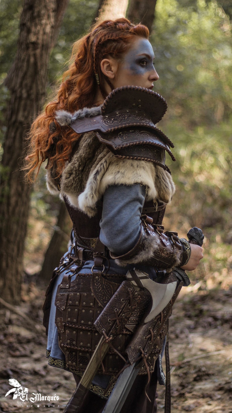 May include: A woman wearing a brown leather armor with fur accents and a sword strapped to her side. She has red hair and blue face paint. The image is taken in a forest setting.