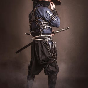 May include: A person wearing a blue and black outfit with a wide-brimmed hat, holding a sword and a variety of knives. The person is standing in a smoky environment.