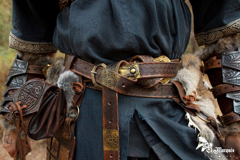 May include: A brown leather belt with intricate gold metal accents and a Celtic knot design. The belt is worn over a dark blue tunic and secured with a large, ornate buckle.