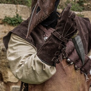 May include: A person wearing a brown leather plague doctor mask with a black tricorn hat and brown leather gloves holding a knife. The person is wearing a brown leather vest and a white shirt. The background is a stone wall.