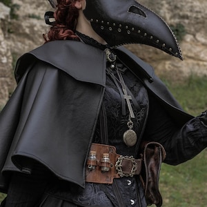 May include: A person wearing a black leather plague doctor mask with a long beak, a black leather hat, a black cape, and a black dress. The person is holding a gun in their right hand. The background is a rocky wall.
